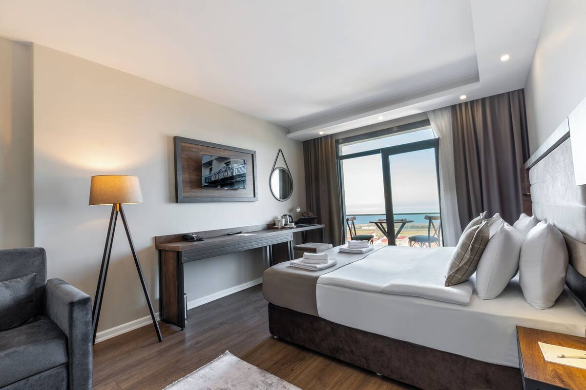 Quarto de hotel moderno com uma cama grande, uma escrivaninha, uma cadeira, um abajur e vista para o mar.