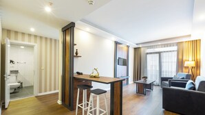 Una sala de estar moderna con un sofá, una mesita de centro y un televisor. Hay una zona de cocina con mostrador y taburetes. El ambiente tiene suelo de madera y papel tapiz a rayas.