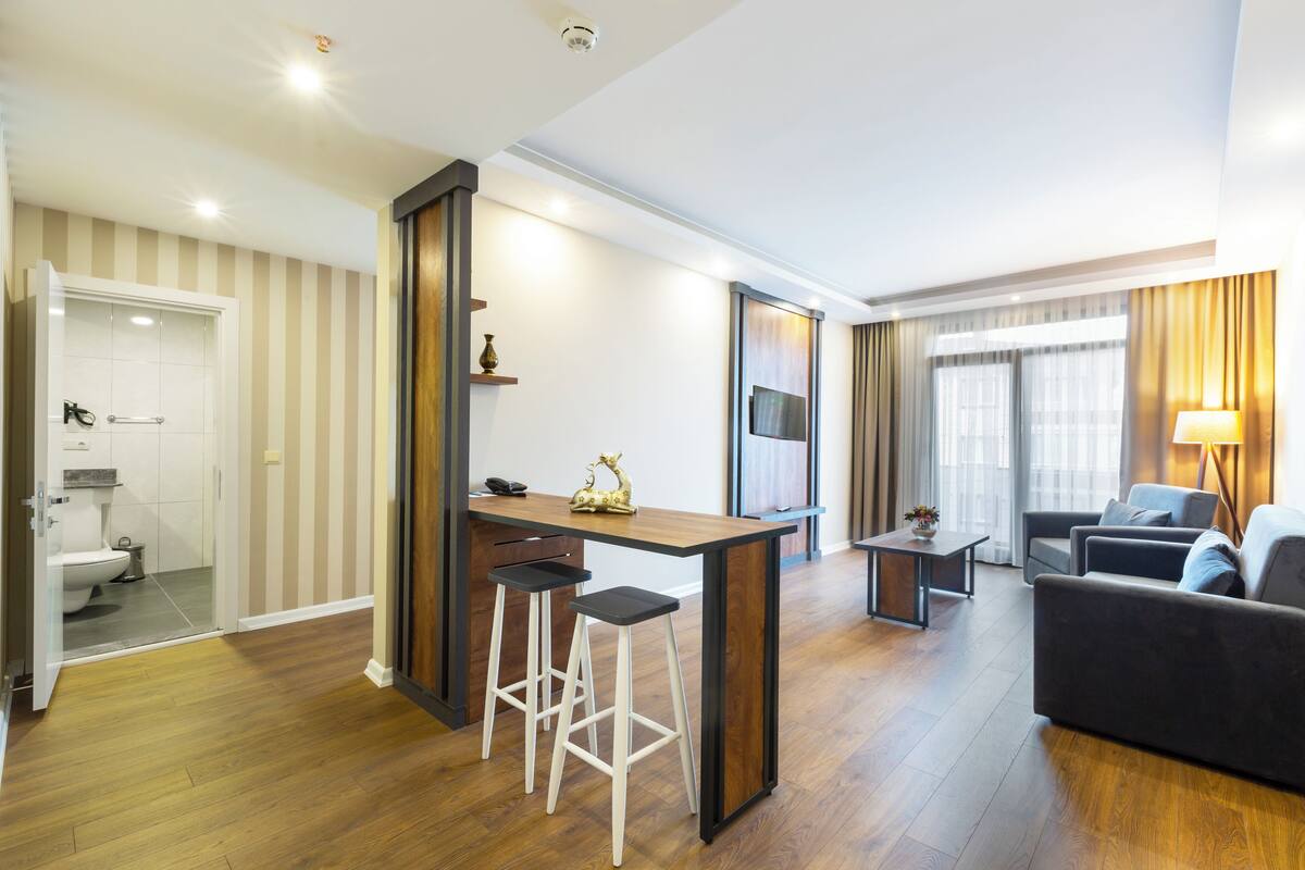 Uma sala de estar moderna com sofá, mesa de centro e televisão. Há uma área de cozinha com bancada e banquetas. O cômodo possui piso de madeira e papel de parede listrado.
