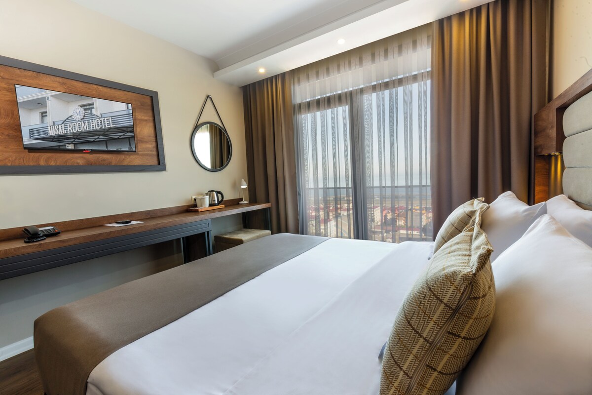 A hotel room with a bed, a desk, a chair, a telephone, a mirror, and a window with curtains.