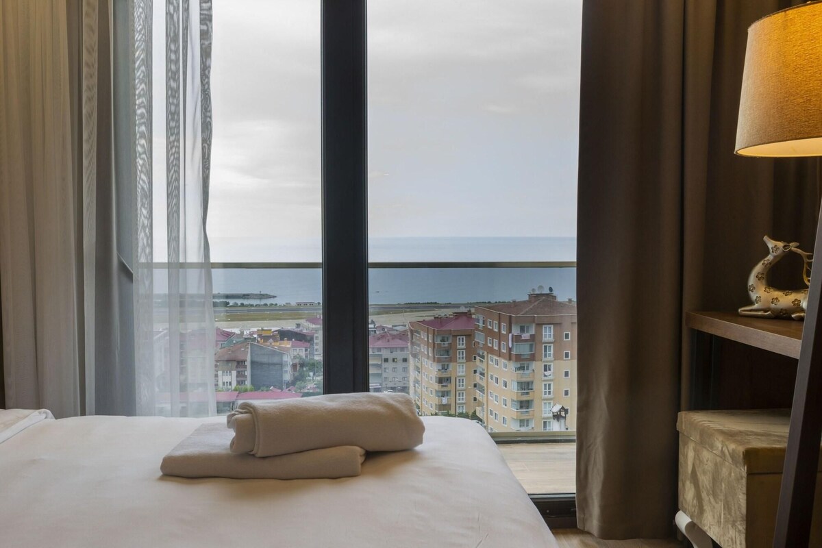 A hotel room with a large window, a bed with white linens, a bedside table with a lamp, and a view of buildings and a body of water.
