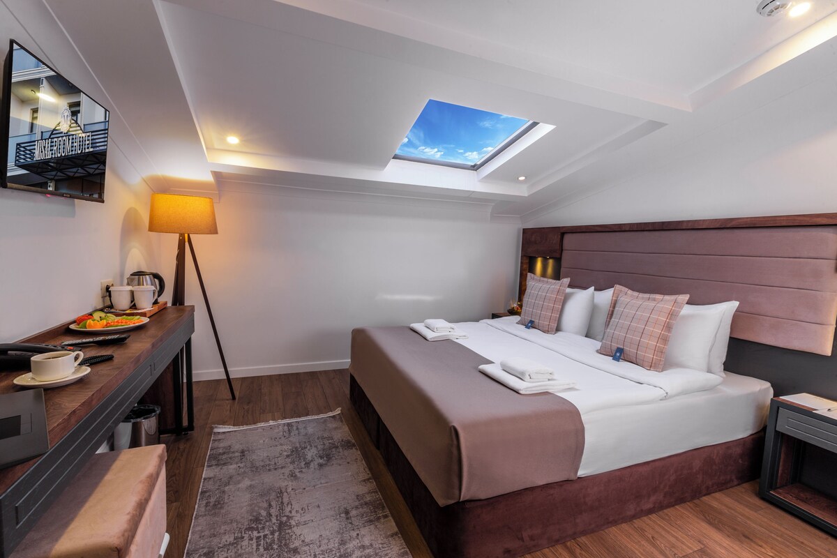 A modern hotel room with a skylight, a bed with multiple pillows, a bedside table with a lamp, and a TV mounted on the wall.