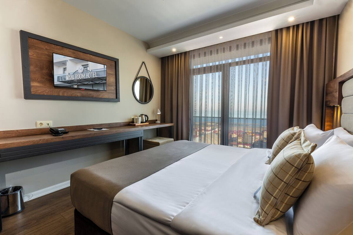 Quarto de hotel com uma cama grande, uma escrivaninha com telefone, um espelho e vista da cidade através de amplas janelas.