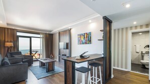 Una sala moderna con sofá, mesa de centro y zona de comedor con taburetes. Hay un ventanal con cortinas, un cuadro en la pared y vista al mar.