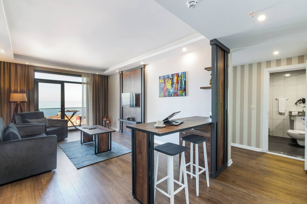 Uma sala de estar moderna com sofá, mesa de centro e área de refeições com banquetas. Há uma janela grande com cortinas, um quadro na parede e vista para o mar.
