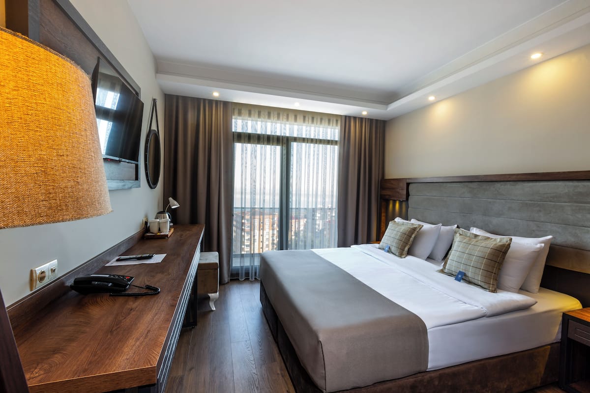 Quarto de hotel com uma cama grande, uma mesa com telefone, televisão e vista para a cidade através de amplas janelas.