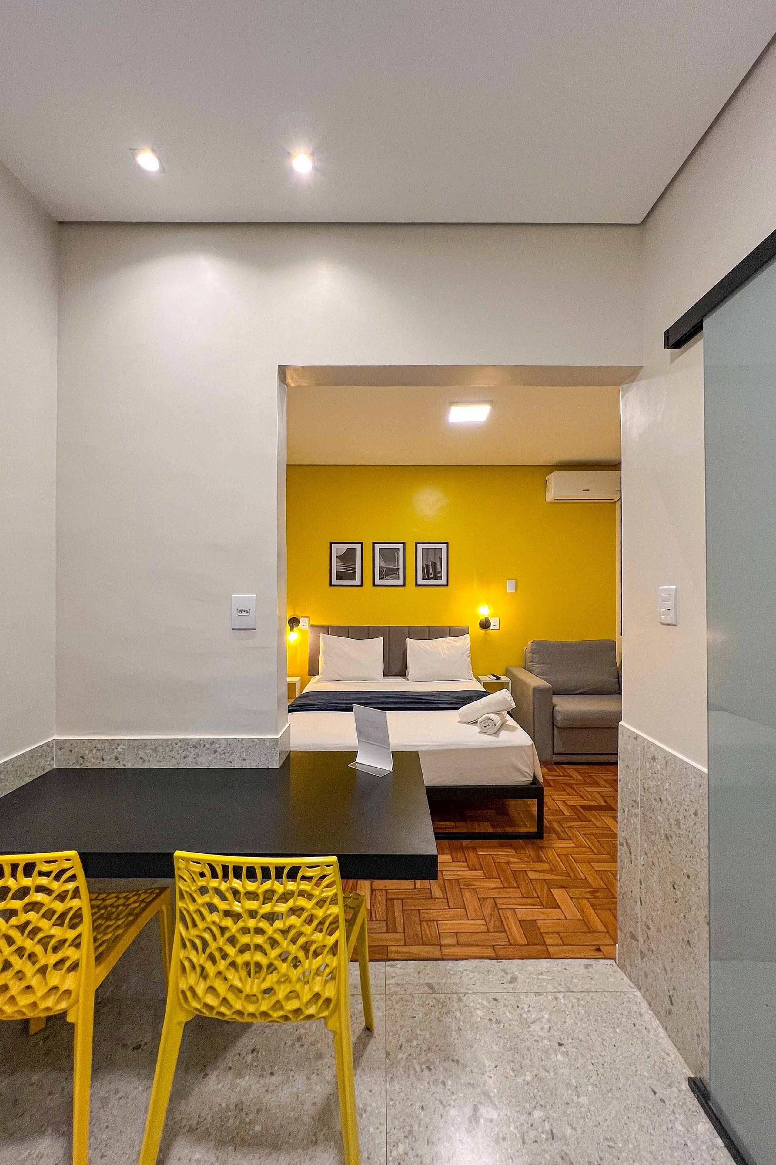 A hotel room with a bed, a desk, and chairs. There is a yellow wall with pictures and a gray sofa.