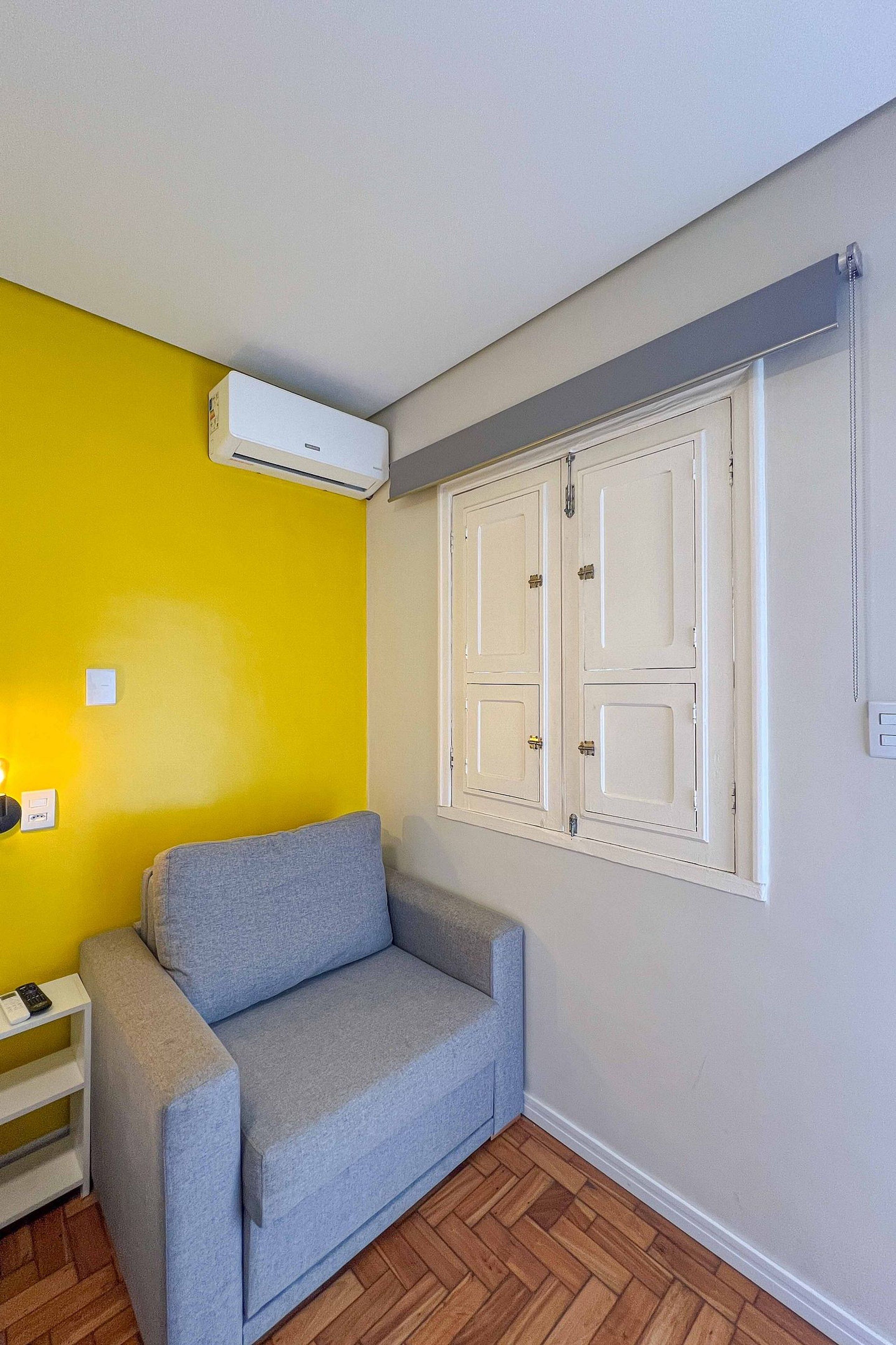 Una habitación con un sofá gris, una pared amarilla, una puerta blanca con un ventanal y un equipo de aire acondicionado instalado en el techo.