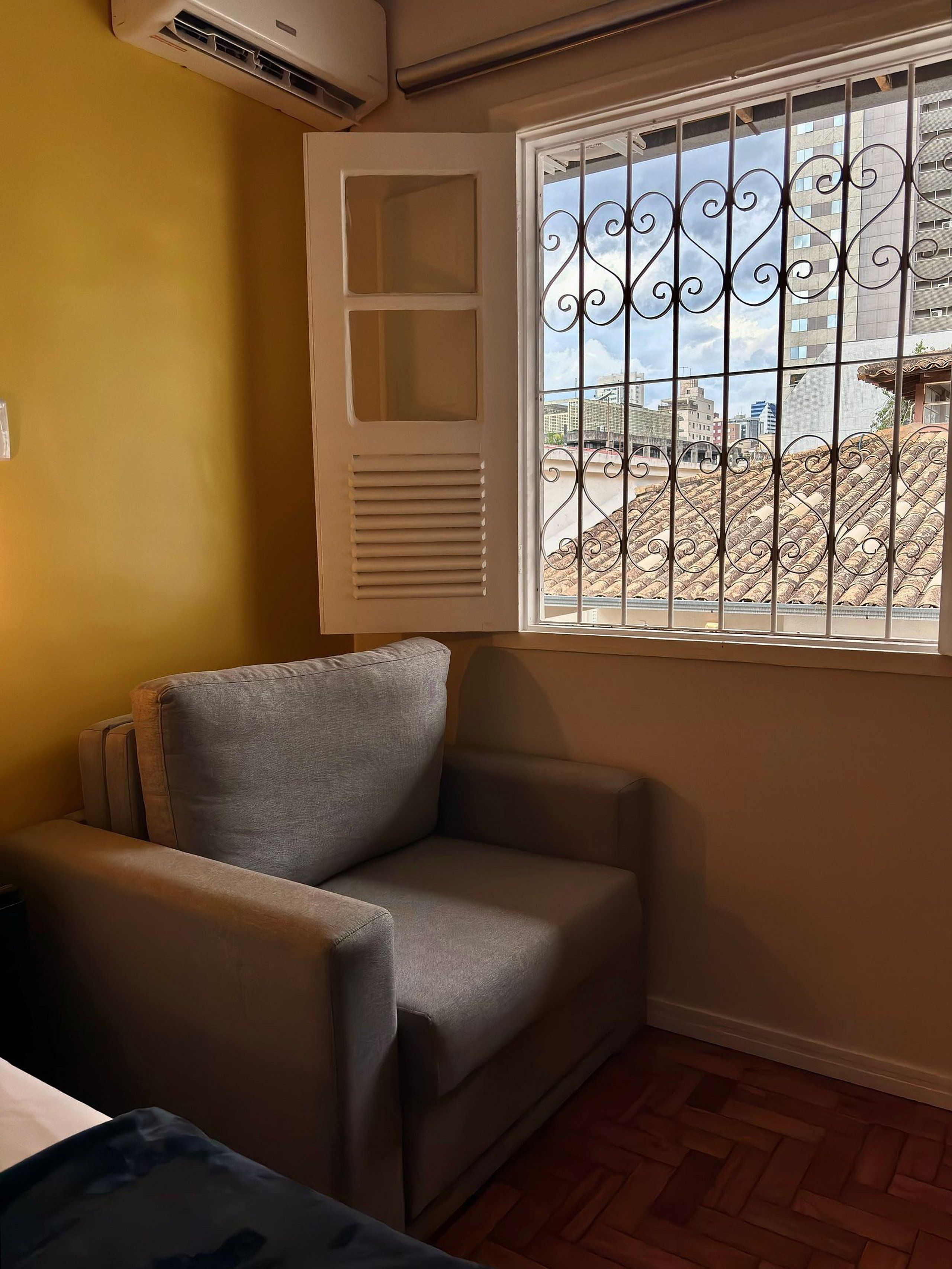 A room with a grey armchair, a window with decorative iron bars, and a view of buildings outside.