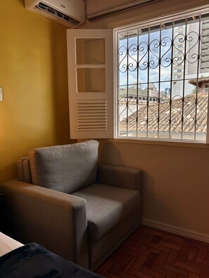 Una habitación con un sillón gris, una ventana con rejas decorativas y vistas a edificios.