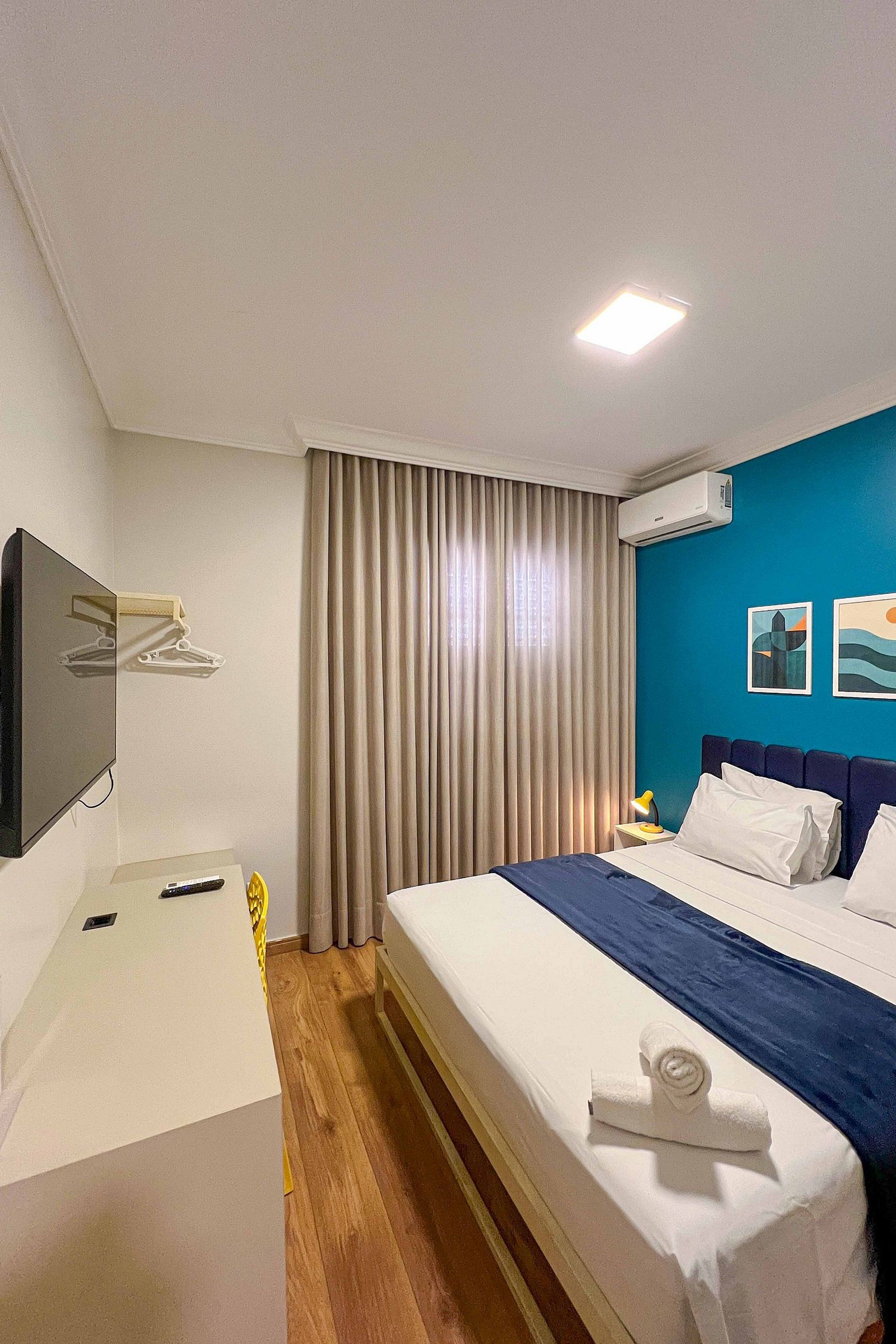 A hotel room with a bed, a desk, a TV, an air conditioner, and a window with curtains.