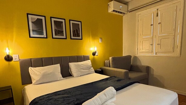 Hotellihuone, jossa on suuri sänky, tuoli, yöpöytä ja seinälle asennettu ilmastointilaite.