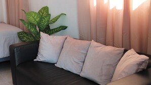 A black leather sofa with four cushions, a large green plant, and a bed with a white duvet in a room with beige curtains. - Live near the Colosseum! Apartment less than 20 min from the airport (San Juan)