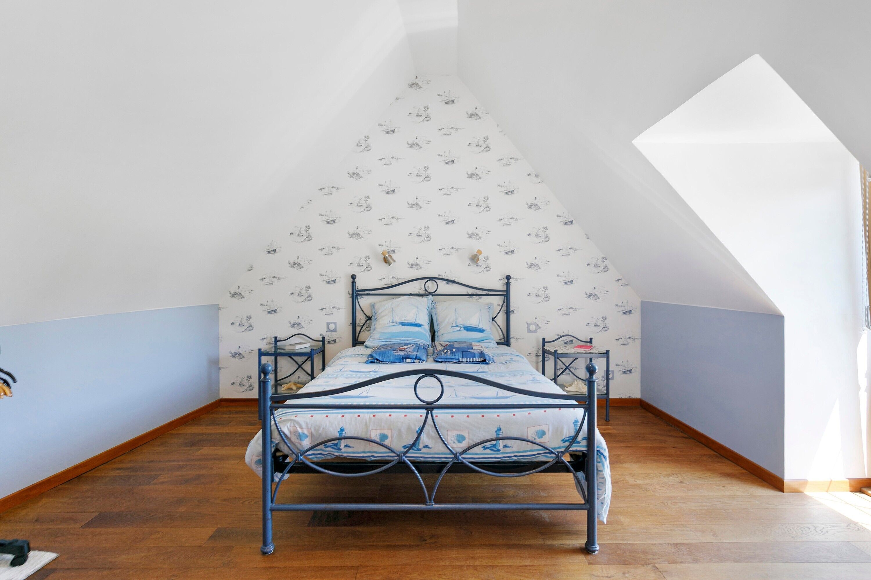 A bedroom with a bed, wooden flooring, a sloped ceiling, and patterned wallpaper.