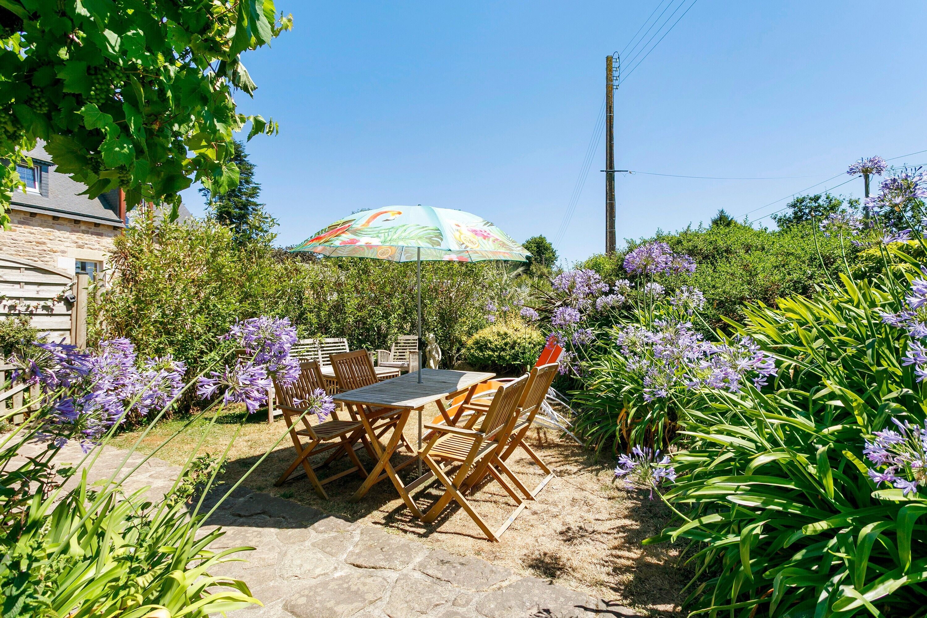 Eine Terrasse im Freien mit Tisch und Stühlen, umgeben von üppigem Grün und violetten Blumen.
