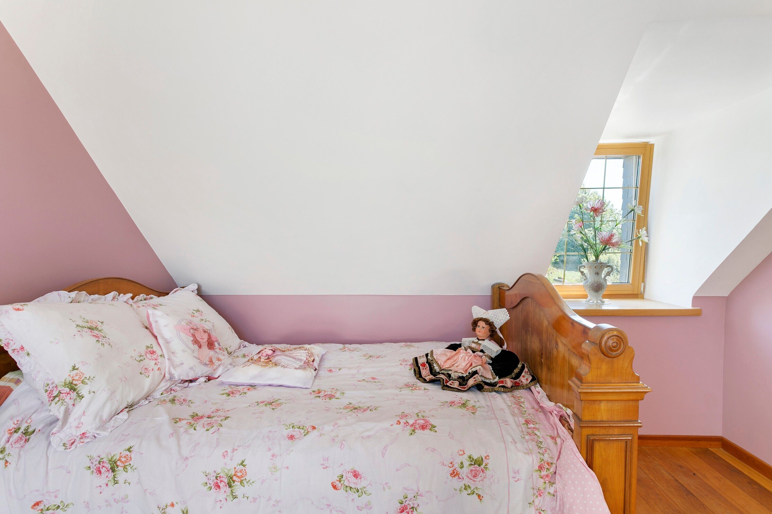 Ein Schlafzimmer mit schrägen Wänden, einem Bettgestell aus Holz und einem Fenster mit Blick auf Grünflächen.