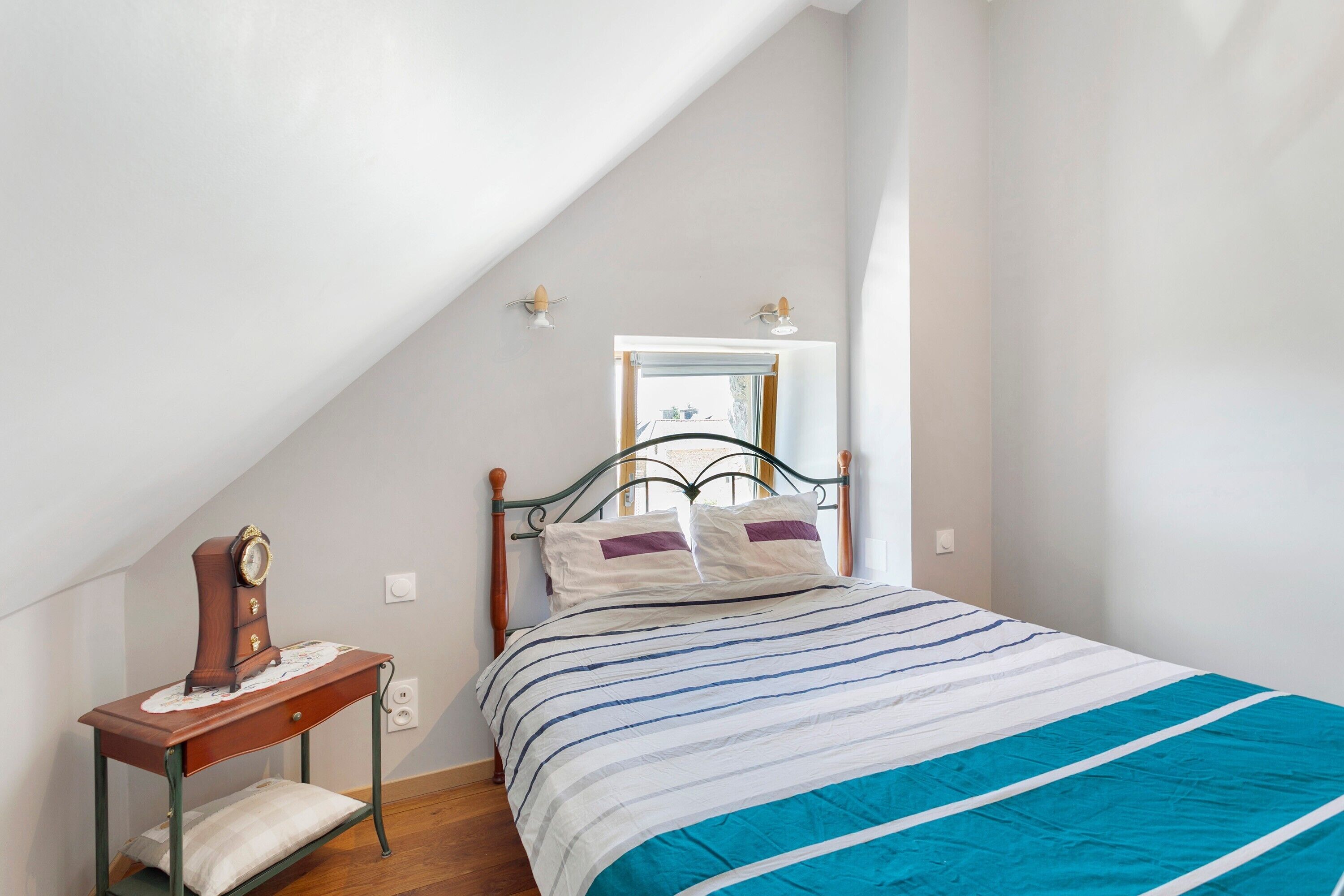 Un dormitorio con techo inclinado, una cama con ropa de cama a rayas, una mesita de noche de madera con un reloj y un espejo en la pared.