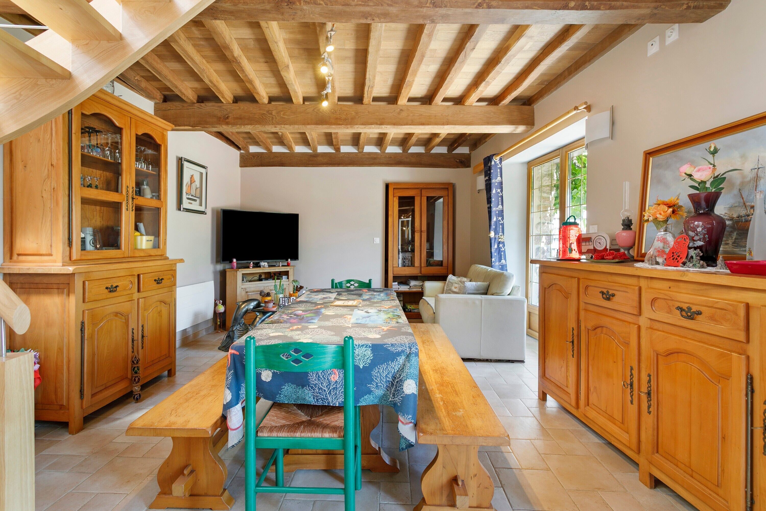 Un coin repas avec une table en bois, des chaises et un téléviseur. On y trouve des meubles de rangement en bois dotés de portes en verre, ainsi qu’une fenêtre avec des rideaux.