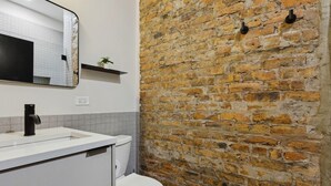 A bathroom with a white sink, black faucet, and a white toilet. There is a mirror above the sink and a brick wall behind it. - Logan Square SRO Hotel (Chicago)