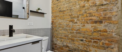 A bathroom with a white sink, black faucet, and a white toilet. There is a mirror above the sink and a brick wall behind it.