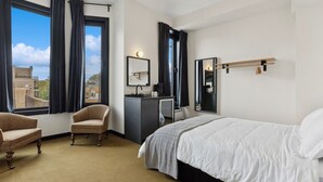 A hotel room with a bed, a desk, a chair, a mirror, and a view of the city. - Logan Square SRO Hotel (Chicago)