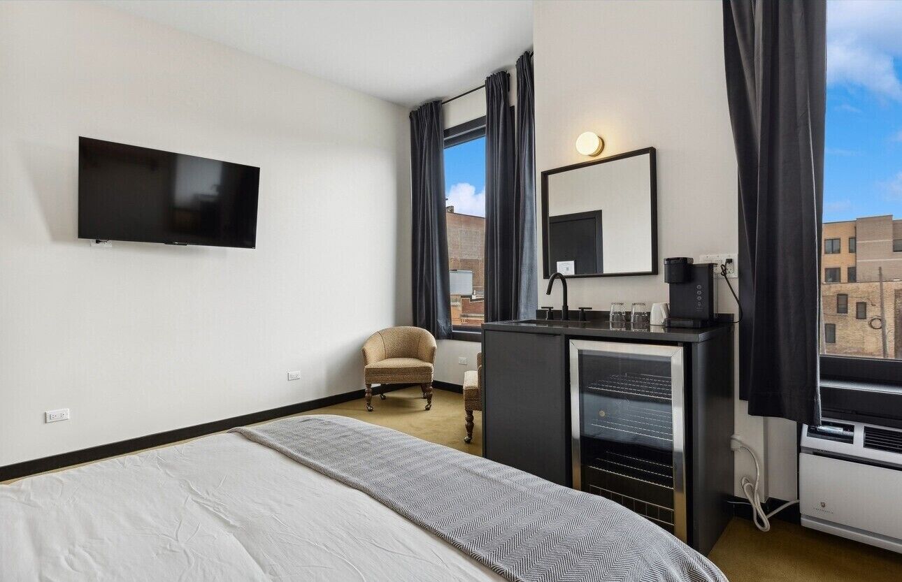 A modern hotel room with a bed, a TV mounted on the wall, a desk with a mirror, and a view of the city through large windows.