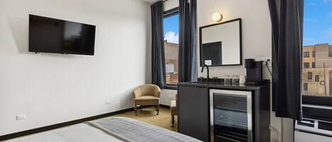 A modern hotel room with a bed, a TV mounted on the wall, a desk with a mirror, and a view of the city through large windows.