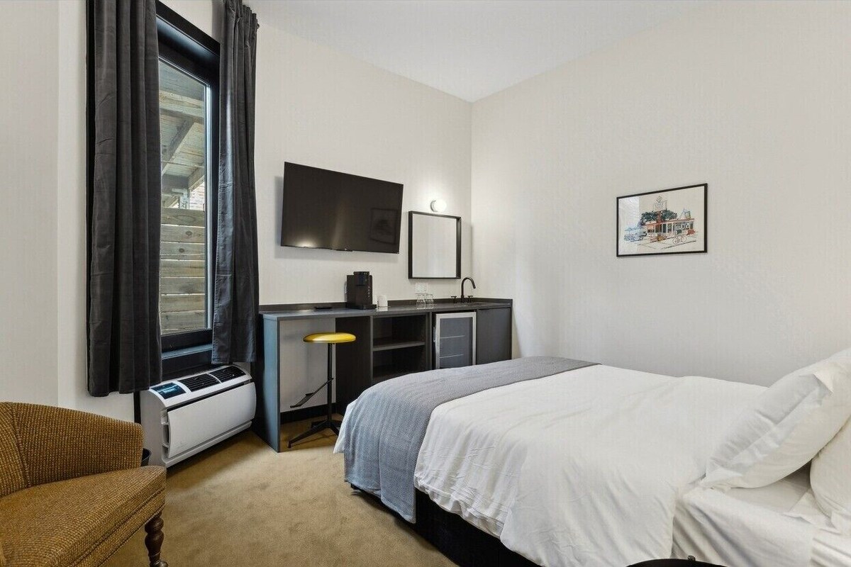 A hotel room with a bed, a desk with a computer, a TV, and a window with curtains.