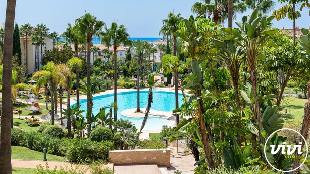 Ein Resort-Pool, umgeben von Palmen und viel Grün, mit Blick auf den Ozean im Hintergrund.