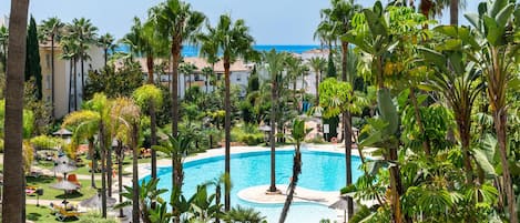A resort pool surrounded by palm trees and greenery, with a view of the ocean in the background.