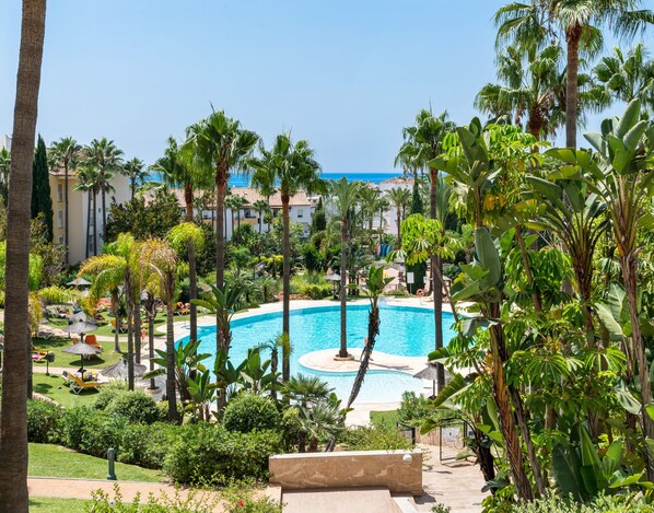 A resort pool surrounded by palm trees and greenery, with a view of the ocean in the background.