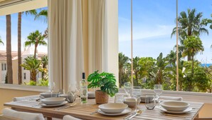 A dining table set with plates, glasses, and a bottle, overlooking a tropical landscape with palm trees and a clear sky.