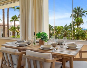 A dining table set with plates, glasses, and a bottle, overlooking a tropical landscape with palm trees and a clear sky.