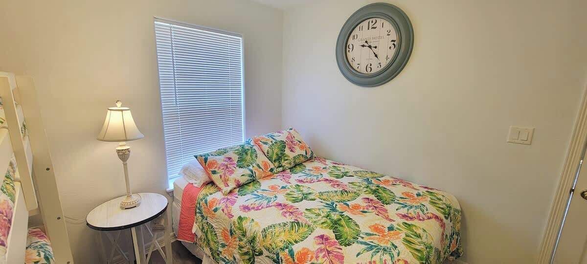 A bedroom with a bed, a round wall clock, a lamp, and a window with blinds.
