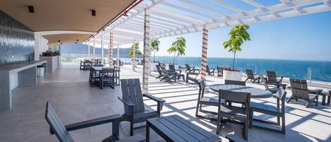 Un espace extérieur moderne aménagé avec des tables et des chaises, un bar et une vue sur l’océan.