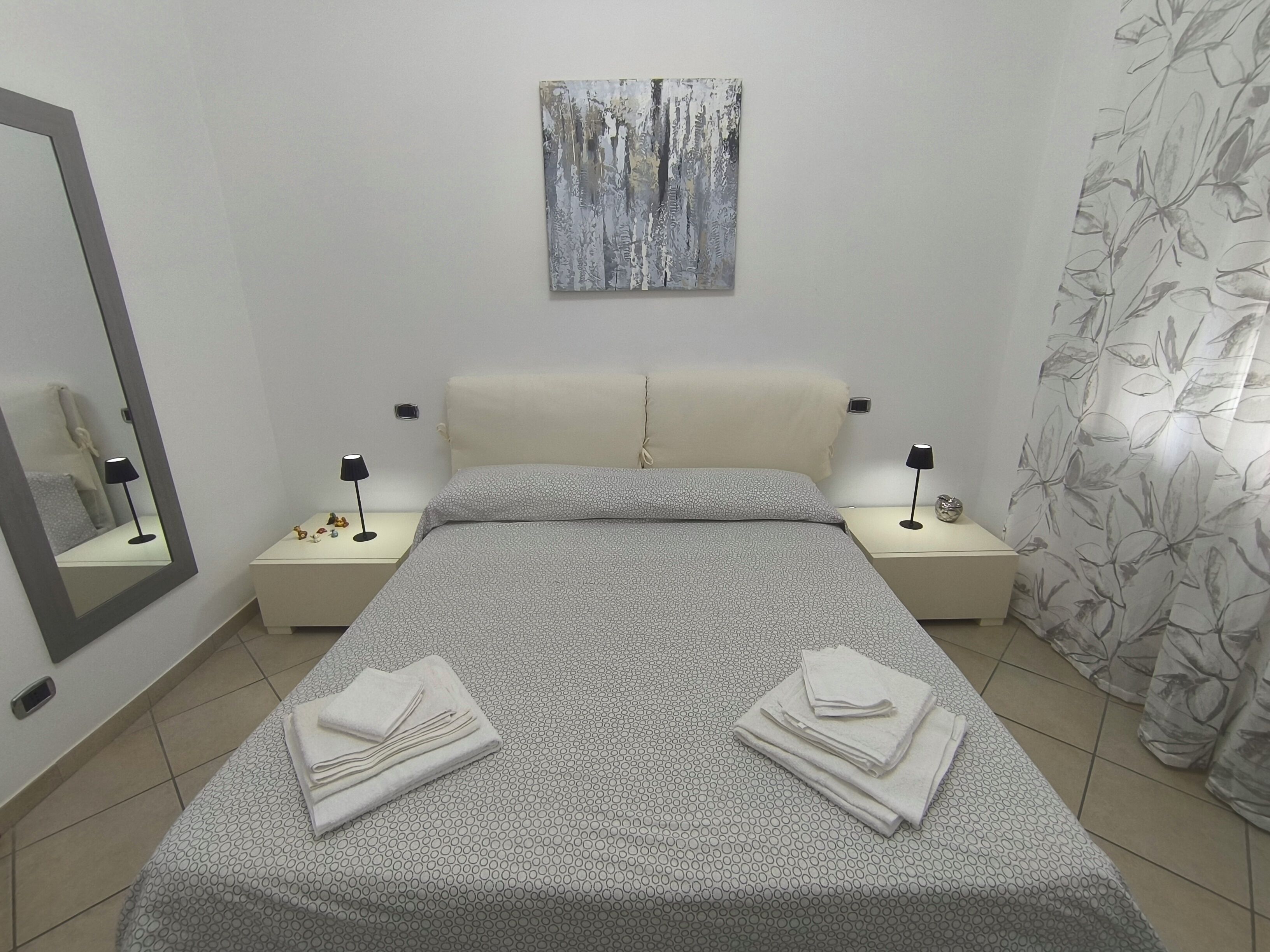 A bedroom with a bed, two bedside tables, a nightstand, a mirror, a painting on the wall, and a curtain.