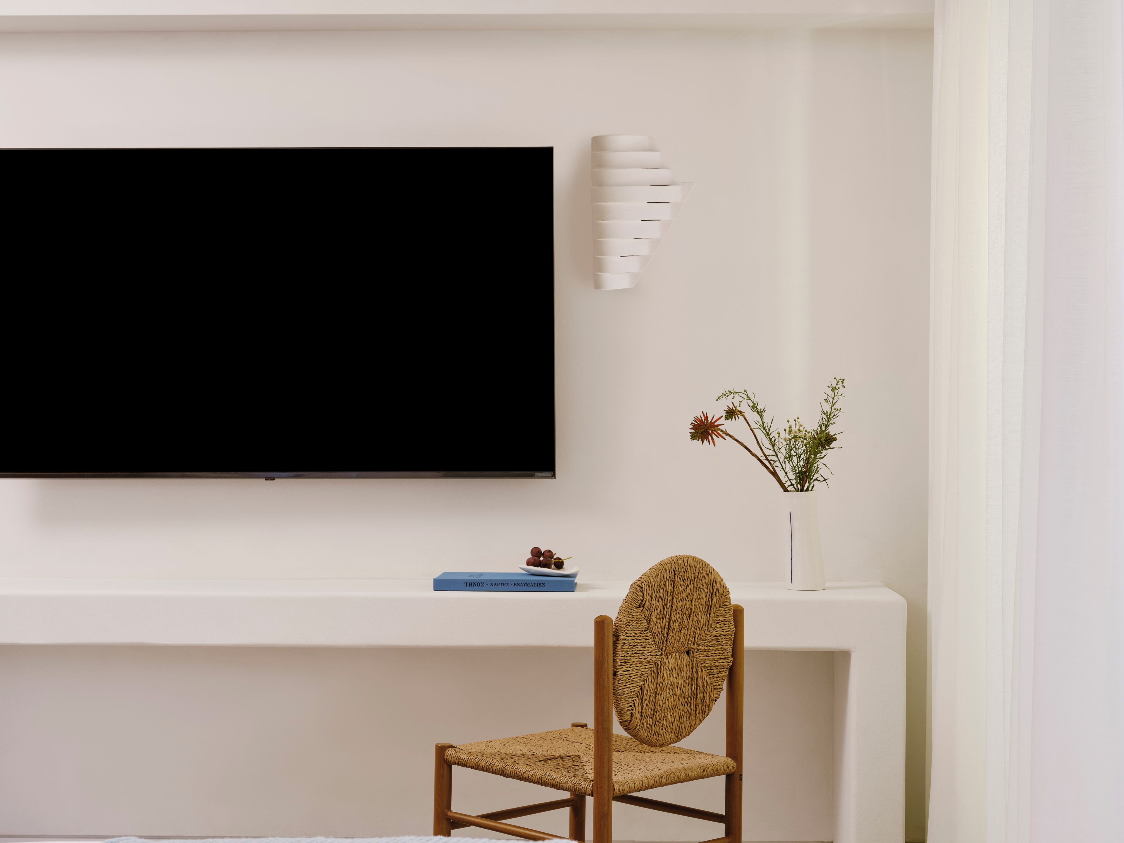 Ein modernes Wohnzimmer mit einem flachbildfernseher an einer weißen Wand, einem gewebten Sessel, einem kleinen weißen Konsolentisch mit einer Vase getrockneter Blumen und einem Buch mit dem Titel 'THINOS - NAPHTAES'.