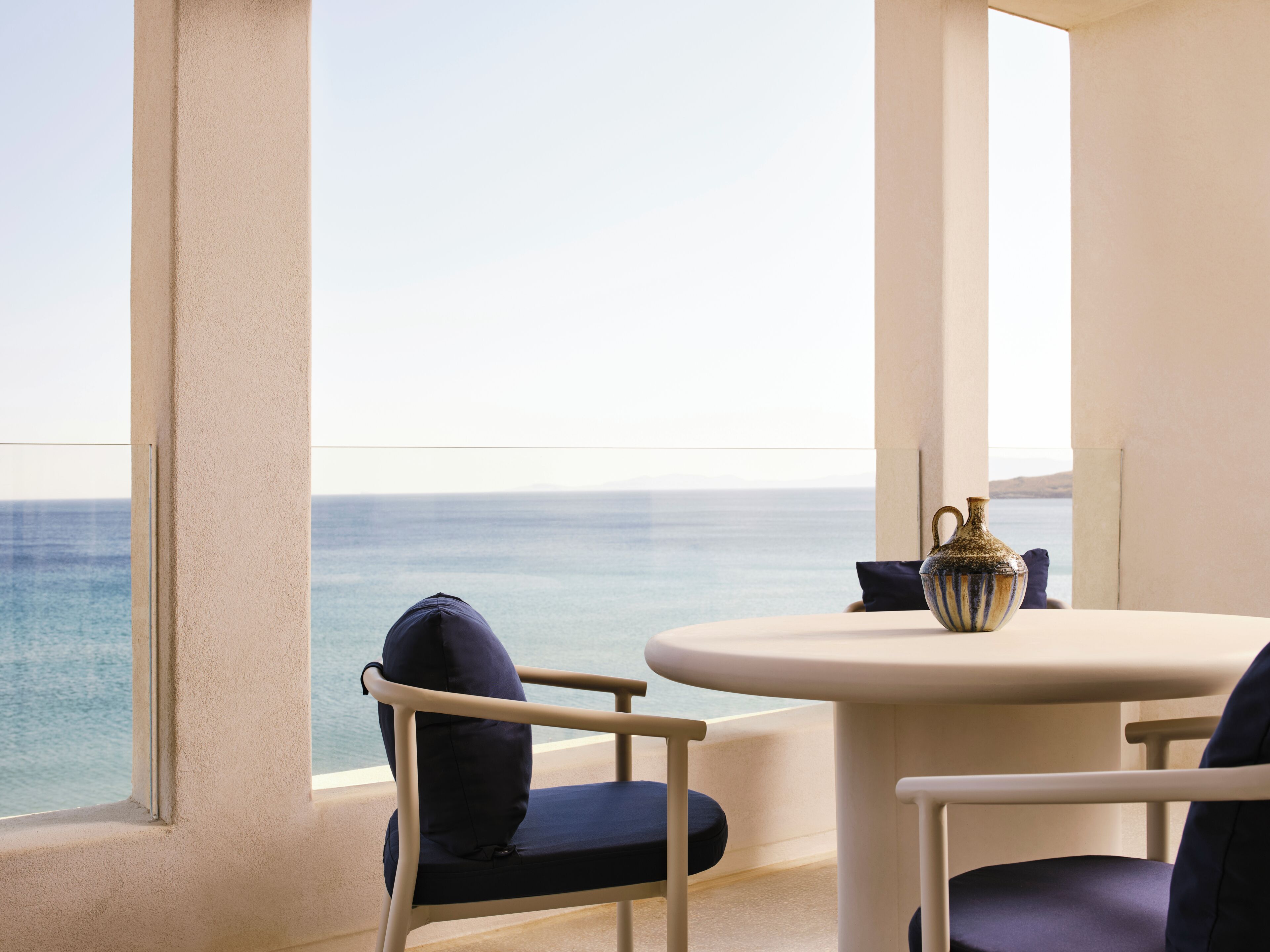 A room with a view of the sea, a round table, two chairs, and a vase.