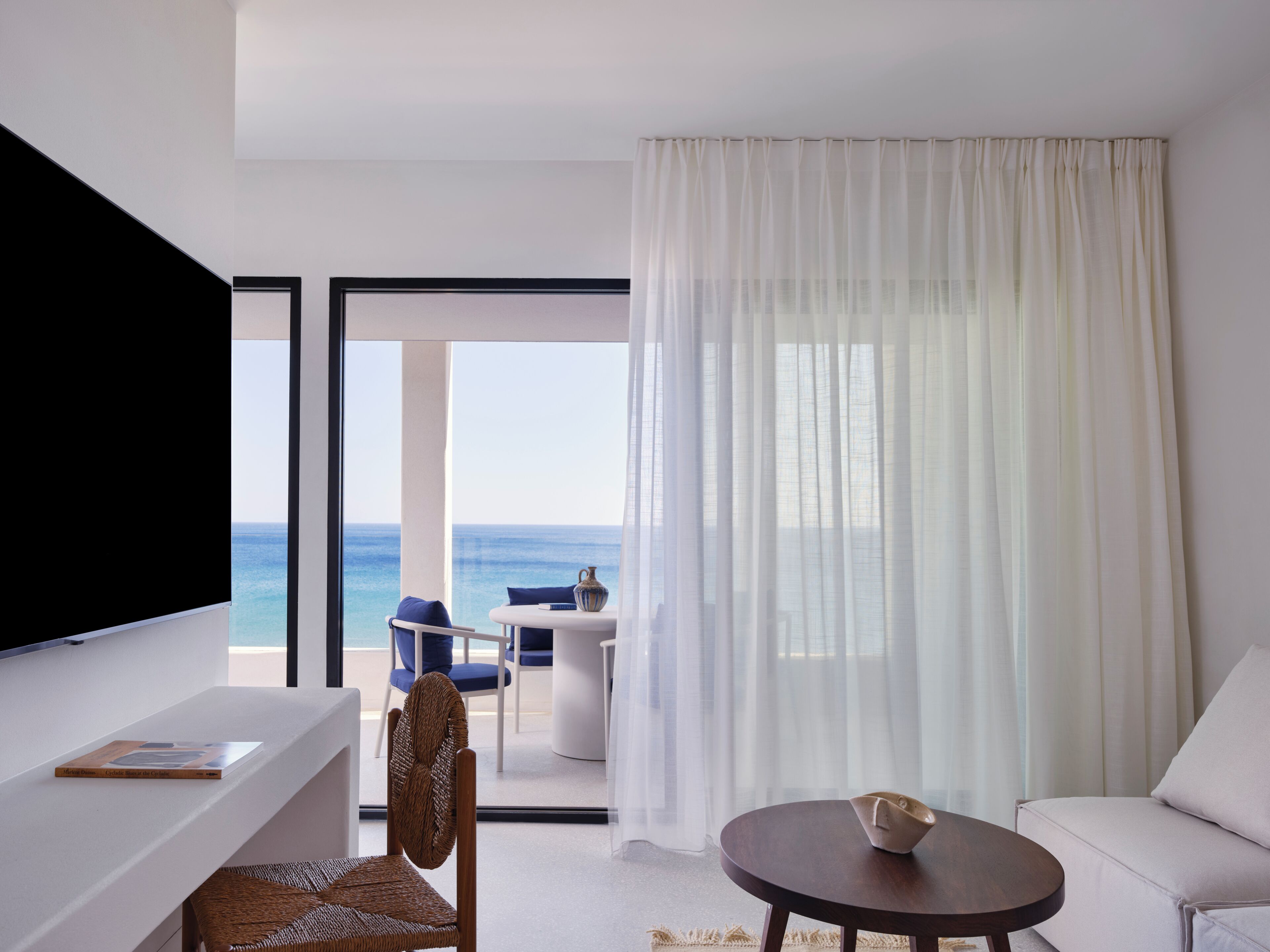 A modern hotel room with a large TV, a dining area with a table and chairs, and a view of the ocean.