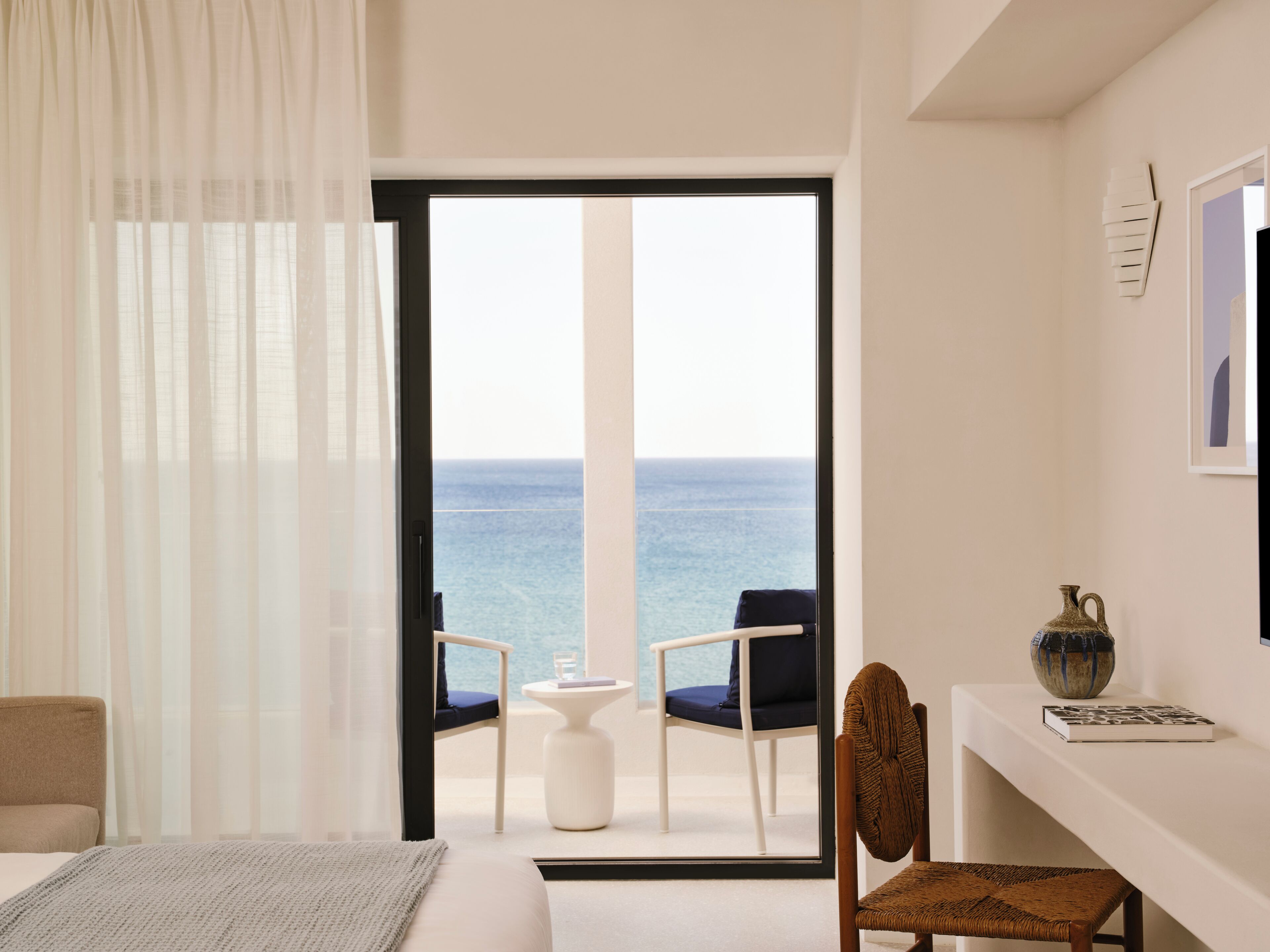 A hotel room with a bed, a chair, a small table, a vase, and a view of the sea.