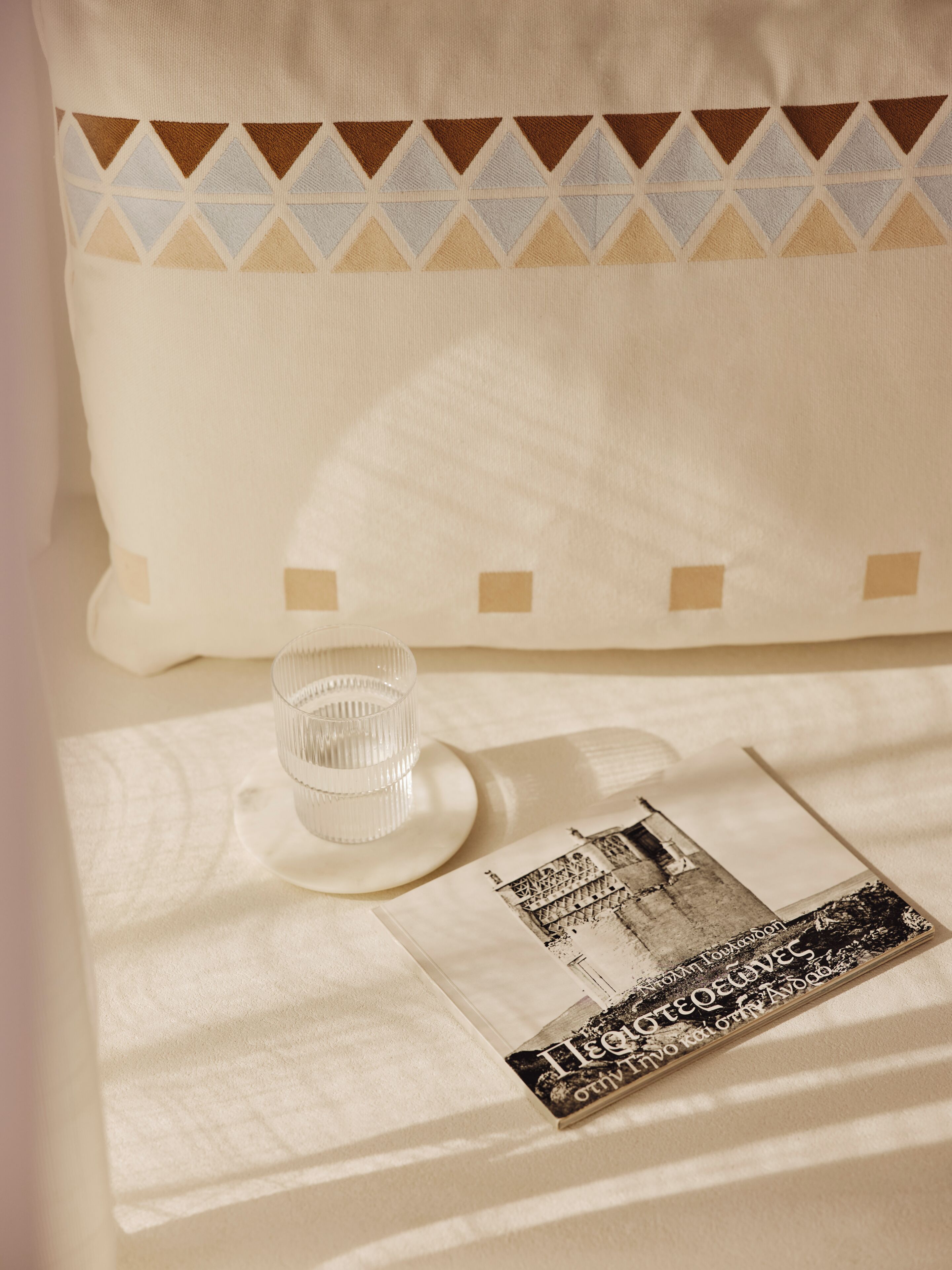 Une chambre d’hôtel bien rangée, avec un oreiller orné d’un motif géométrique, un verre transparent posé sur une assiette blanche et une brochure imprimée sur le lit.