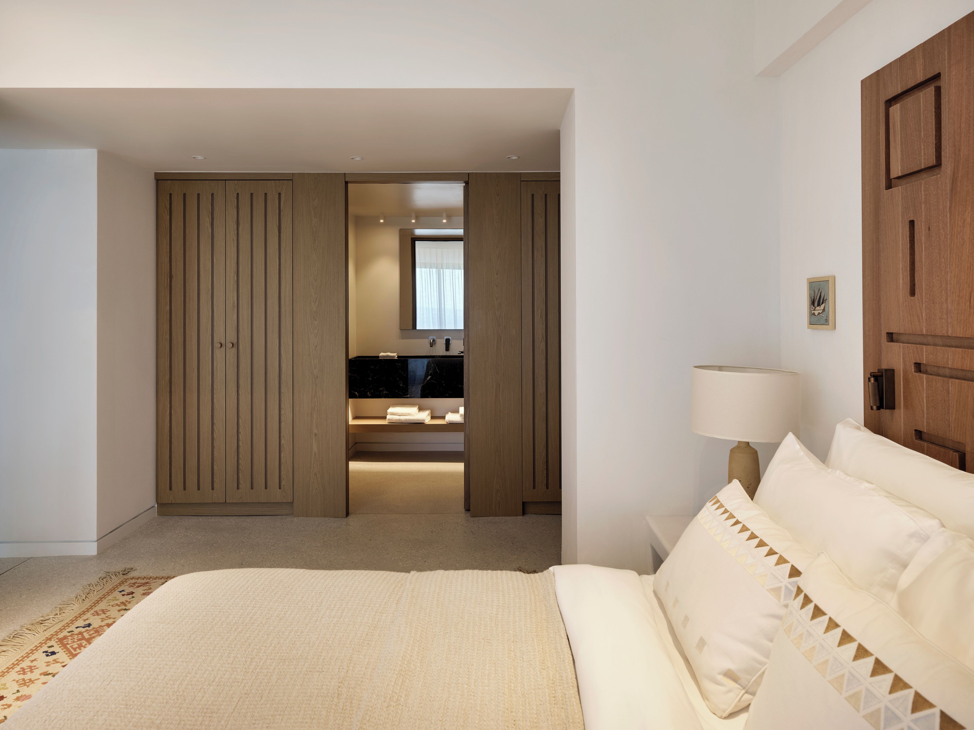 A bedroom with a large bed, a nightstand with a lamp, a wooden wardrobe, and a bathroom with a mirror and sink.