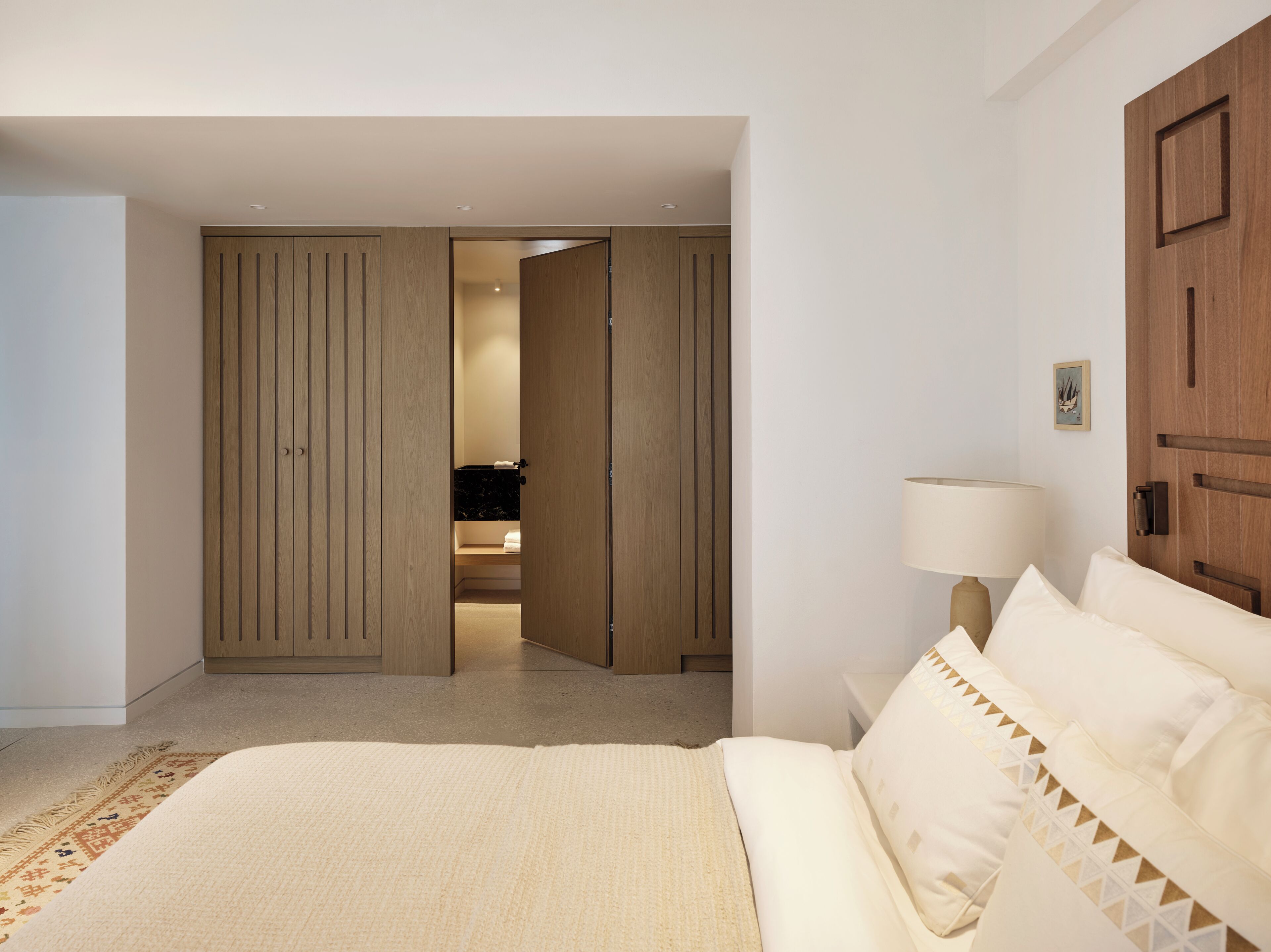 Een slaapkamer met een bed, nachtkastje, lamp, houten garderobe en een badkamer die door een open deur zichtbaar is.