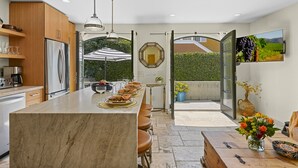 A modern kitchen with a marble island, stainless steel appliances, and a dining area with a view of the garden. - Casa al Mare - Boho Mission Beach Retreat w/ A/C (San Diego)