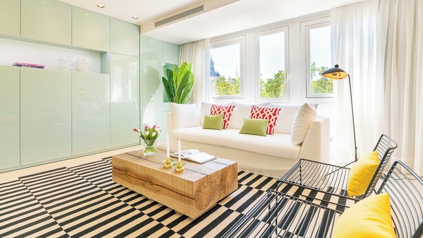 Un salón moderno con un sofá blanco, una mesa de centro de madera, una alfombra a rayas negras y blancas, y un ventanal con cortinas.