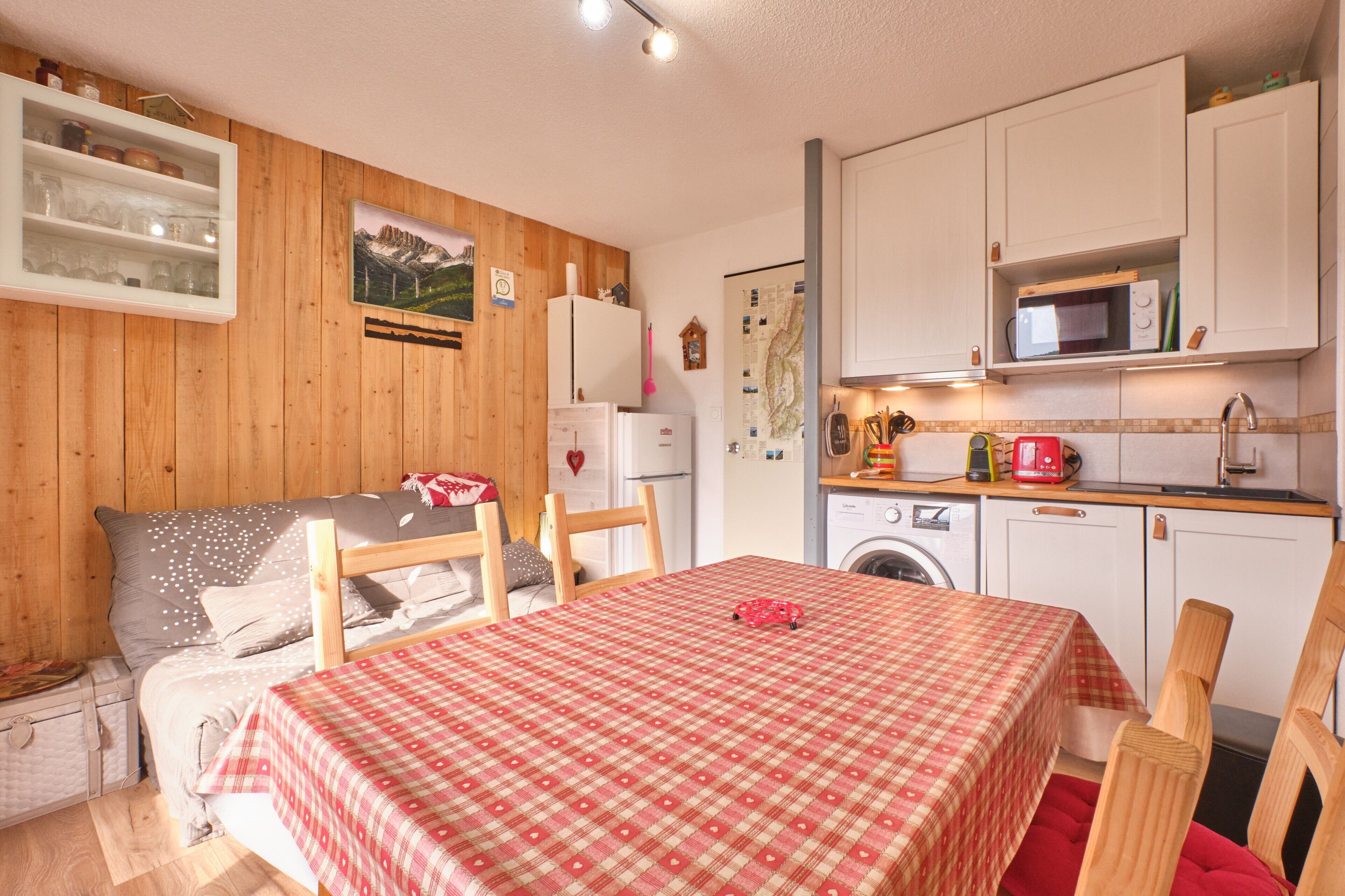 Une pièce confortable avec un lit, une table recouverte d’une nappe à carreaux, une chaise en bois, un lave-linge et un coin cuisine équipé de placards et d’un four à micro-ondes.