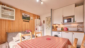 A cozy room with a bed, a table with a checkered tablecloth, a wooden chair, a washing machine, and a kitchen area with cabinets and a microwave.