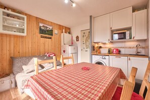 A cozy room with a bed, a table with a checkered tablecloth, a wooden chair, a washing machine, and a kitchen area with cabinets and a microwave.