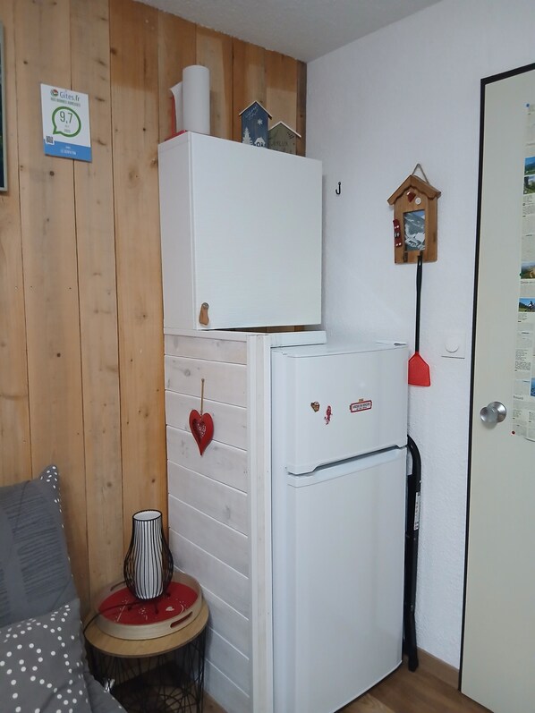 A compact room with a white refrigerator, a wooden wall, a small table with a lamp, and a door with a calendar. - 'Le Serpaton' apartment with mountain view and balcony (Gresse-en-Vercors)