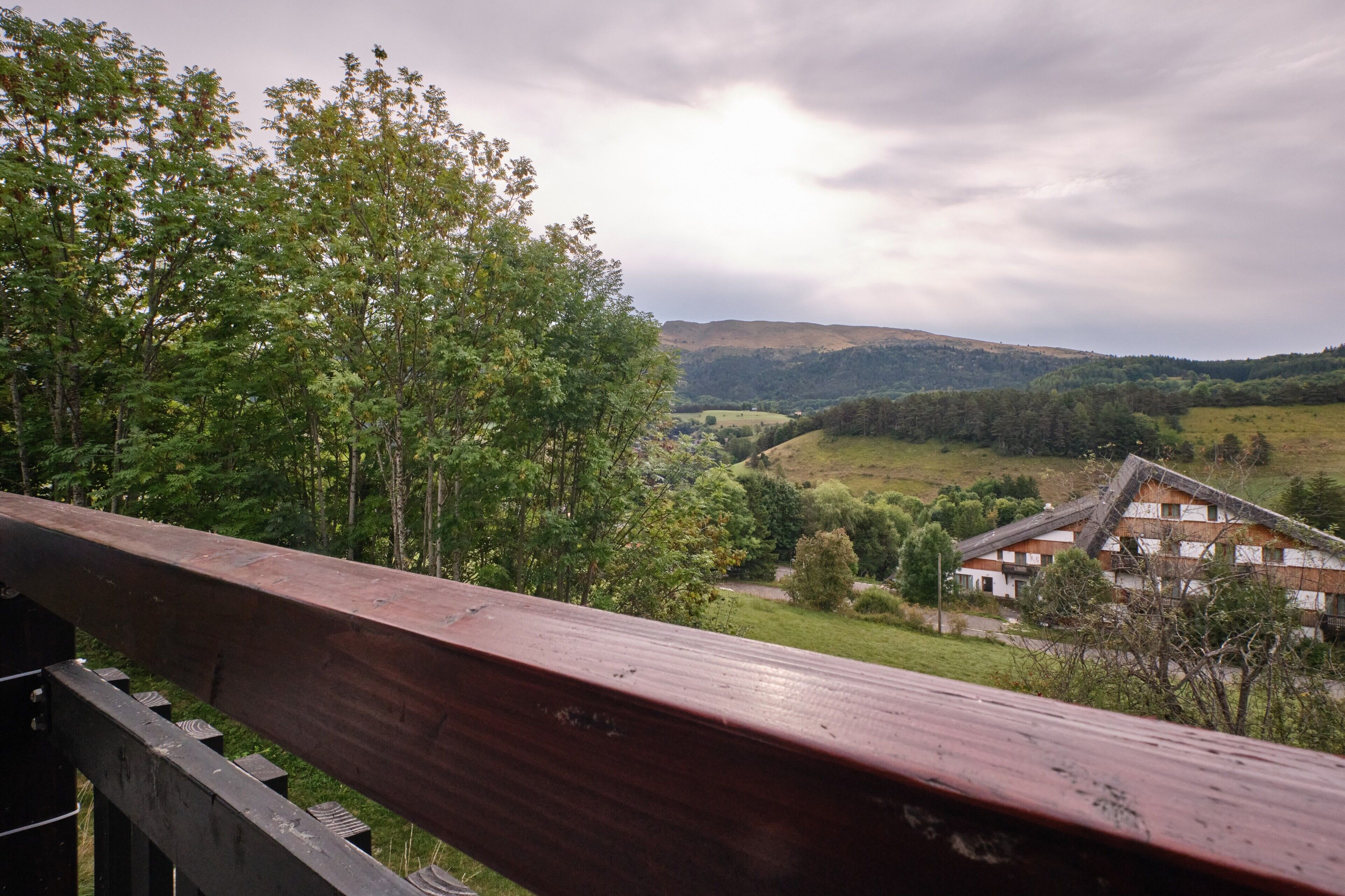 Vista de uma paisagem rural com uma ponte de madeira, uma casa com telhado inclinado e vegetação ao redor.